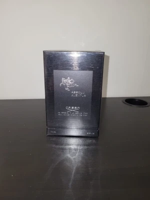 Creed Absolu Aventus parfym 75 ml - Lyxig parfym från Creed, modellen Absolu Aventus. Flaskan rymmer 75 ml och kommer i en elegant svart förpackning med silverdetaljer och Creed-logga. Kartongen har en exklusiv känsla och är signerad på toppen. Perfekt för dig som gillar exklusiva dofter.