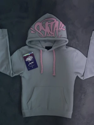 Grå hoodie med rosa snöre från synaworld  - Snygg grå hoodie från synaworld med rosa graffiti-tryck på huvan och matchande rosa dragsnören. 80% bomull 20% polyester. Top kvalitet.