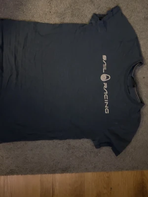 Mörkblå Sail Racing t-shirt - Mörkblå t-shirt från Sail Racing med vitt tryck på bröstet. Klassisk rund halsringning och korta ärmar. Tillverkad i mjukt bomullsmaterial för en skön känsla. Perfekt för en avslappnad och sportig stil.