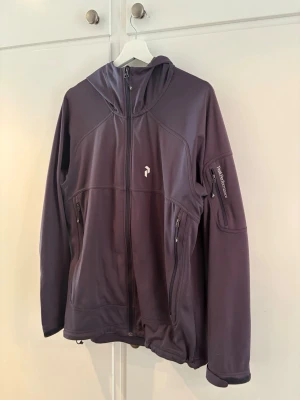 Grå Softshell från Peak Performance - Snygg grå softshell från Peak Performance med huva och dragkedja framtill. Jackan har flera praktiska fickor med dragkedja, inklusive en på ärmen. Perfekt för blåsiga dagar och outdoor-aktiviteter.