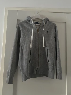 Grå hoodie från Polo Ralph Lauren - Snygg grå hoodie från Polo Ralph Lauren med dragkedja, vita dragsnören och klassisk blå logga på bröstet. Hoodien har två fickor framtill och ribbade muddar vid ärmar och nederkant. Perfekt för en chill och stilren look.