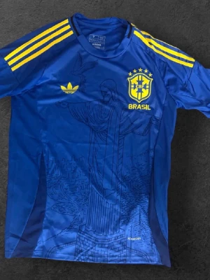  Brasilien fotbollströja Adidas - Snygg blå fotbollströja från Adidas med Brasilien-tryck och gula detaljer. Tröjan har gula ränder på axlarna, CBF-logga och Adidas-logga på bröstet samt ett stort tryck av Kristusstatyn på framsidan. Perfekt för fotbollsfans!