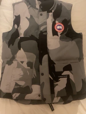 Camouflage dunväst från Canada Goose - Snygg dunväst från Canada Goose i grått, svart och vitt camomönster. Västen har två stora fickor framtill, tryckknappar och klassisk Canada Goose-logga på bröstet. Perfekt för lager-på-lager och streetwear-stil. Materialet är dunfyllt för extra värme. Har använt den ett fåtal gånger
