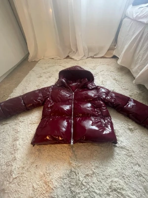Vinröd glansig jacka från Nelly använd en gång. - Snygg vinröd jacka med glansig finish och luva. Jackan har breda kanaler, dragkedja framtill och är riktigt fluffig. Perfekt för kalla vinterdagar och ger en cool streetwear-look. Materialet är syntet med en blank yta.