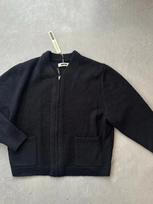 Svart kofta med dragkedja från Woodbird - Stilren svart kofta från Woodbird med dragkedja framtill och två praktiska fickor. Koftan har ribbade muddar och är tillverkad i ett mjukt stickat material. Perfekt för lager-på-lager och enkel att matcha med olika outfits. Köpt på Vallgatan 12 i Göteborg för några månader sedan. Använd någon gång. Nytt skick med prislappen kvar. 