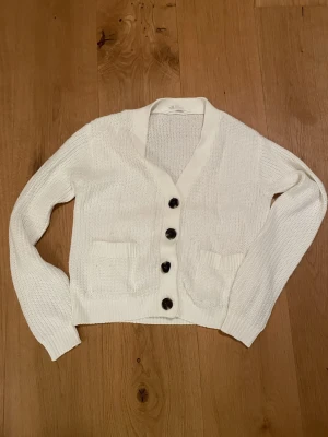 Vit stickad kofta med knappar - Snygg vit stickad kofta med fyra stora svarta knappar framtill och två små fickor. Koftan har lång ärm, ribbade muddar och en djup v-ringning. Perfekt för lager på lager och enkel att matcha med olika outfits.