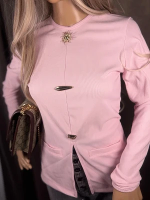 Ljusrosa ribbad blus med detaljer - Trendig ljusrosa blus i ribbat material med långa ärmar och unika metallknappar framtill. Rund halsringning och dekorativ brosch upptill. Blusen har två fickor framtill och en liten slits nedtill. Perfekt för dig som gillar stilrena men ändå unika plagg.