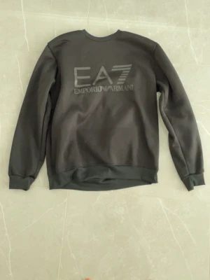 Svart EA7 Emporio Armani sweatshirt - Snygg svart sweatshirt från EA7 Emporio Armani med stor logga på bröstet. Tröjan har rund halsringning, långa ärmar och ribbade muddar. Perfekt för en stilren och sportig look.