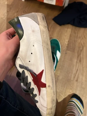 Golden Goose vita sneakers med röd stjärna - Golden Goose sneakers i vitt läder med grön häl och grå detaljer. Sidan pryds av en röd stjärna och skorna har grå skosnören. Yttersulan är beige med ett slitet utseende och undersidan är i gummi med fiskbensmönster. Perfekt för dig som gillar streetstyle och unika detaljer.