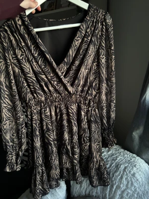 Klänning med djurmönster - Snygg kort klänning från Holly’s med zebramönster i svart och beige. Klänningen har v-ringning, samt lite volangdetaljer nertill. Passar XS-S