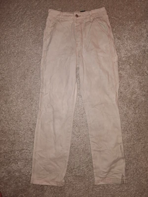 Beiga chinos med raka ben - Säljer ett par beiga jeans med raka ben och flera fickor, perfekta för en avslappnad och stilren look. Jeansen har synliga sömmar och klassisk knappgylf. Materialet känns mjukt och bekvämt, passar till många olika outfits. Storlek ca 38EU. Plagget har synlig missfärgning på baksidan. (Se bild 2 och 3).