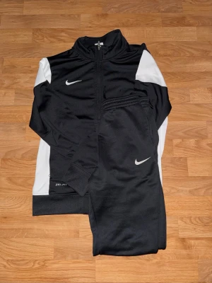 Svart och vit Nike Dri-FIT träningsset - Nike Dri-FIT träningsset med svart och vit jacka och matchande svarta byxor. Jackan har dragkedja och vita paneler på ärmarna, medan byxorna har en liten vit Nike-logga. Perfekt för sport och träning, tillverkad i polyester för maximal komfort.