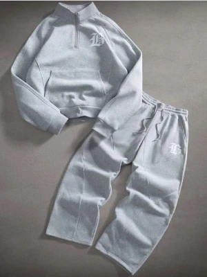 Grått loungewear-set med tryck - Mysigt grått loungewear-set med matchande tröja och byxor. Tröjan har halv dragkedja, hög krage och magficka, medan byxorna har resår i midjan. Båda plaggen har ett vitt tryckt bokstavsmotiv på bröst och ben. Perfekt för chill dagar hemma.