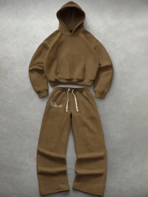 Brunt loungewear-set med hoodie - Mysigt loungewear-set i brun färg med oversized hoodie och matchande byxor. Hoodien har stor ficka framtill och byxorna har dragsko i midjan samt broderad text på ena benet. Perfekt för chill dagar hemma.