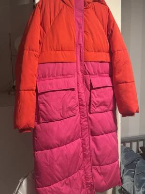 Jacka ifrån YAS. Storlek S (oversize, jag är eg. large) - Säljer en lång, oversized dunjacka i tvåfärgat blockmönster med rött upptill och rosa nedtill. Jackan har huva, stora fickor framtill och dragkedja. Perfekt för kalla vinterdagar och ger en riktigt cool look.