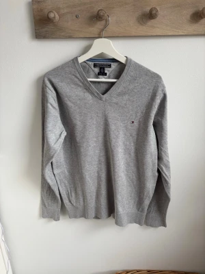 Grå V-ringad tröja Tommy Hilfiger - Snygg grå V-ringad tröja från Tommy Hilfiger med liten broderad logga på bröstet. Tillverkad i mjukt material och har ribbade muddar vid ärmslut och nederkant. Perfekt för en clean och stilren look. Finns lite ytludd bak, se bild 5.