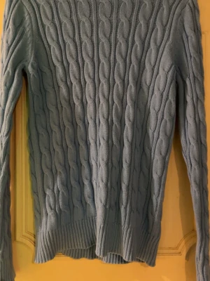 Ljusblå kabelstickad tröja - Snygg ljusblå kabelstickad tröja med rund hals och långa ärmar. Tröjan har ribbade muddar vid ärmslut och nederkant, vilket ger en klassisk och stilren look. Perfekt att matcha med jeans eller kjol för en avslappnad vibe. Aldrig använd