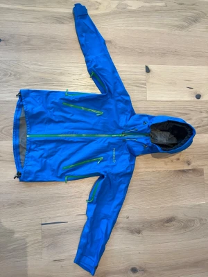 Blå vindjacka barn Gore-Tex från NORRØNA - Blå vindjacka i Gore-Tex för barn från NORRØNA med gröna dragkedjor och huva. Jackan har flera praktiska fickor och är perfekt för blåsiga dagar och outdoor-aktiviteter.