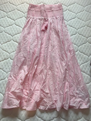 Ljusrosa boho-kjol med tofsar - Söt ljusrosa kjol i bohostil med smock i midjan och dekorativa tofsar framtill. Kjolen har ett diskret mönster och är gjord i ett tunt, luftigt tyg som känns perfekt för sommaren. Passar dig som gillar en avslappnad och romantisk look.