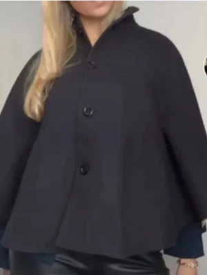 Svart poncho från H&M - Stilren svart cape från H&M med knappar framtill och hög krage. Modellen är vid och har fickor på sidorna samt slät insida. Perfekt för lager-på-lager och ger en cool siluett till din outfit. 