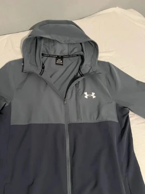 Gråblå vindjacka från Under Armour - tja en riktig skön vindjacka i storlek (S) skicken på den är 10/10! absolut inget fel på den verkan skada eller nån typ av fläck.  Materialet är lätt och känns skönt att ha på sig den💯🙌🏻
