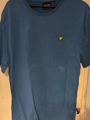 Blå t-shirt från Lyle & Scott - Snygg blå t-shirt från Lyle & Scott med klassisk rund hals och det ikoniska gula logobroderiet på bröstet. Perfekt basic-plagg i mjuk bomull som funkar till allt. Enkel och stilren design som passar till jeans eller shorts. Notera fläcken på bild 3 som ej går bort.