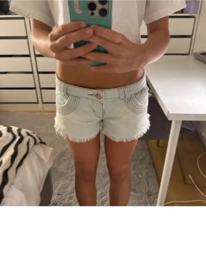 Ljusblå jeansshorts med fransig kant - Snygga ljusblå jeansshorts med fransig kant och dekorativa sömmar på fickorna. Klassisk femficksmodell med knapp och dragkedja framtill. Perfekta för varma dagar.