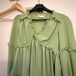 Grön volangblus från AWARE - Snygg ljusgrön blus från AWARE med volangdetaljer längs ringningen och över bröstet. Blusen har v-ringning och långa ärmar, tillverkad i ett mjukt, lätt tyg som ger ett flowy intryck. Perfekt för dig som gillar romantiska detaljer.