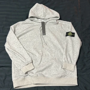Grå hoodie från Stone Island - Snygg ljusgrå hoodie från Stone Island med klassisk logopatch på vänster ärm. Hoodien har huva med dragsko, ribbade muddar och en avslappnad passform. Perfekt för dig som gillar streetwear och vill ha något stilrent men ändå ikoniskt. Helt ny.