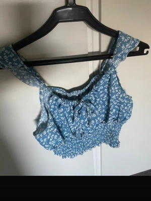 Blommig blå croppad linnetopp - Söt blå croppad linnetopp med vitt blommönster och breda axelband. Toppen har smockad nederkant och en liten knytning framtill för extra detalj. Perfekt för varma dagar och ger en fräsch sommarkänsla.