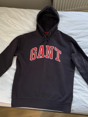 Svart hoodie från GANT med tryck - Snygg svart hoodie från GANT med stor röd och vit logga på bröstet. Klassisk huva med dragsko och ribbade muddar vid ärmar och nederkant. Perfekt för en chill och sportig look. Tillverkad i mjukt bomullsmaterial.