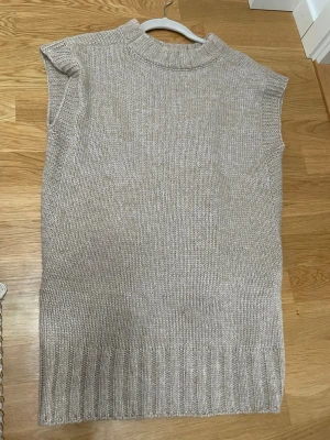 Beige stickad väst med slits - Snygg beige stickad väst med ribbad kant och slits i sidorna. Perfekt att slänga över en skjorta eller t-shirt för en chill och trendig look. Västen har en högre halsringning och är i en loose passform.
