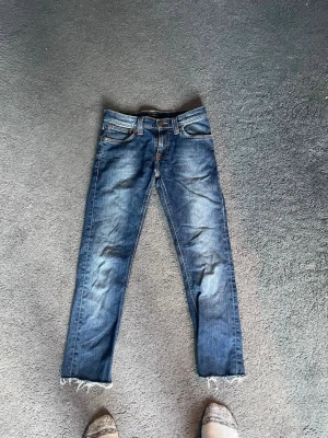 Blå jeans från Nudie Jeans - Säljer ett par klassiska blå jeans från Nudie Jeans med snygg slitning och råa, fransiga benslut. Jeansen har fem fickor, orange sömmar och en skön, rak passform. Tillverkade i denim av bomull. Perfekta för en avslappnad och trendig look.