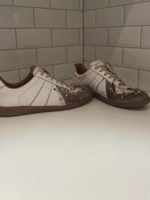 Maison margiela skor - Unika vita maison margiela sneakers med bruna och svarta färgstänk, klassisk låg modell med bruna sulor och vita snören. Snygg retrostil och coolt mönster som sticker ut. Perfekta för dig som vill ha något annorlunda och trendigt. Hör av dig vid frågor eller priset!