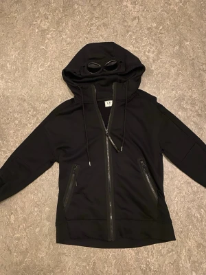 Svart hoodie med glasögon från C.P. Company - Säljer en svart hoodie från C.P. Company med hel dragkedja, två snedställda fickor med dragkedja och ikoniska inbyggda glasögon i huvan. Hoodien har justerbara snören och är tillverkad i mjukt material. Perfekt för dig som vill sticka ut med en unik detalj.