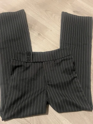 Kostymbyxor  - Grå/svarta utsvängda kostymbyxor med vita ränder. Har resår i midjan och knappar