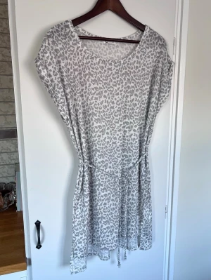 Grå/vit leopardklänning med knytning i midjan från Cubus, storlek M - Försvinner sen 31:a mars om den inte blir såld innan dess :) Grå/vit klänning med leopardmönster och knytband i midjan. Klänningen har rund halsringning och är kortärmad, lite uppvikt vid ärmarna (syns på bild nr 6). Mycket sparsamt använd!