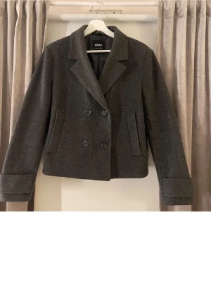 Mörkgrå kort kappa från ONLY - Stilren mörkgrå kort kappa från ONLY med dubbelknäppning och bred krage. Rocken har två snedställda fickor framtill och är tillverkad i ett ull-liknande material. Perfekt för dig som gillar klassisk och trendig look. Storlek M men mer som S (jag själv är en S o passa mig perfekt) säljer för att den inte kommer till användning.