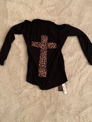 Svart offshoulder topp med leopardkors - Svart långärmad topp från Romwe med ett stort kors i leopardmönster på framsidan. Toppens material känns mjukt och stretchigt, perfekt för en edgy look.