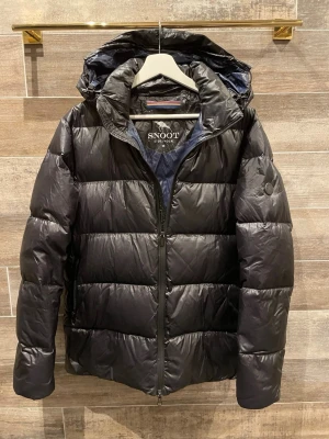Snoot jacka  - Snygg svart dunjacka från Snoot Stockholm med huva och dragkedja framtill. Jackan har en quiltad design och diskret logga på ärmen. Perfekt för kalla vinterdagar och håller dig varm med sin vadderade insida.