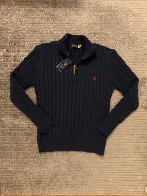 Mörkblå kabelstickad tröja Polo Ralph Lauren - Mörkblå kabelstickad tröja från Polo Ralph Lauren med halv dragkedja och läderdetalj. Tröjan har lång ärm, ribbade muddar och röd broderad logga på bröstet. Perfekt för lager-på-lager och klassisk stil.