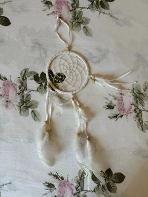 Vit drömfångare med fjädrar - Säljer en stilren vit drömfångare med träpärlor och mjuka vita fjädrar. Ringen är insvept i vitt band och nätet är knutet i ett klassiskt mönster. Perfekt som dekoration till väskan, rummet eller som personlig accessoar.