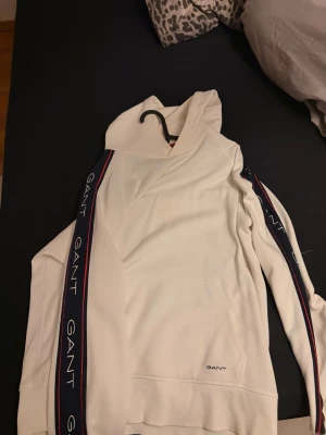 Vit hoodie från GANT med logoband - Snygg vit hoodie från GANT med huva och känguruficka. Längs ärmarna går marinblå och röda band med GANT-loggan tryckt i vitt. Tillverkad i mjukt bomullsmaterial och har ribbade muddar vid ärmslut och nederkant. Perfekt för en avslappnad och trendig look.
