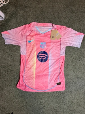 Rosa FC Barcelona matchtröja Nike - Snygg rosa FC Barcelona matchtröja från Nike med klubbmärke och Spotify-logga på bröstet. Tröjan har korta ärmar, V-ringning med ljusblå kant och detaljer i gult och blått. Tillverkad i lätt och ventilerande funktionsmaterial, perfekt för fotboll eller träning.