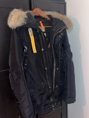 Svart Parajumpers parkas med pälskrage - Svart parkajacka från Parajumpers med ikonisk gul rem och detaljer i metall. Jackan har en stor huva med fluffig pälskant, flera fickor och patch på ärmen. Perfekt för kalla dagar och riktigt snygg streetstil.