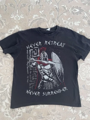 Svart t-shirt med spartansk krigare - Cool svart t-shirt med tryck av en spartansk krigare, röd detalj på hjälmen och texten 'Never Retreat Never Surrender' framtill. Klassisk passform och rund hals. Perfekt för dig som gillar unika och tuffa motiv.