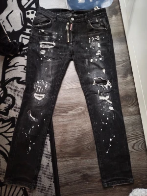Dsq2 Svarta slitna jeans med färgstänk - Säljer ett par svarta jeans med skinny fit, slitningar och hål på benen samt vita färgstänk för en edgy look. Jeansen har klassisk femficksmodell och dragkedja med knapp. Perfekta för dig som gillar streetstyle och vill sticka ut. Är dsq2 storlek 