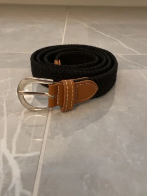 Svart flätat bälte med skinn-detaljer - Stilrent svart flätat bälte med bruna skinn-detaljer och silverspänne. Perfekt accessoar för att lyfta din outfit med en klassisk touch. Bältet har snygga kontrastsömmar och är flexibelt i materialet.