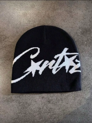 Svart mössa från Corteiz med vit logga - Snygg svart mössa från Corteiz med stor vit logga i graffiti-stil framtill. Mjuk och stretchig passform, perfekt för att hålla värmen och ge en cool streetwear-look. Passar dig som gillar trendiga accessoarer och vill sticka ut.