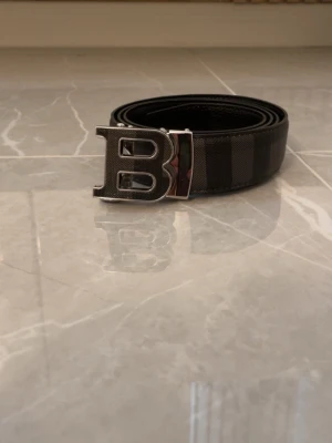 Svart bälte med B-spänne - Stilrent svart bälte från Balmain med ett stort, silverfärgat B-spänne. Bältet har diskreta ränder och är tillverkat i skinn med en elegant finish. Perfekt accessoar för att lyfta din outfit.